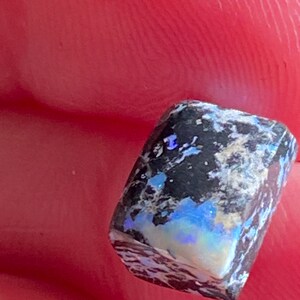 May include: A close-up of a rough-cut opal gemstone. The stone has a dark, speckled surface with flashes of blue, white, and iridescent colors. The opal is held between fingers, showing its natural, unpolished state.