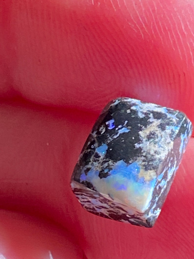 May include: A close-up of a rough-cut opal gemstone. The stone has a dark, speckled surface with flashes of blue, white, and iridescent colors. The opal is held between fingers, showing its natural, unpolished state.