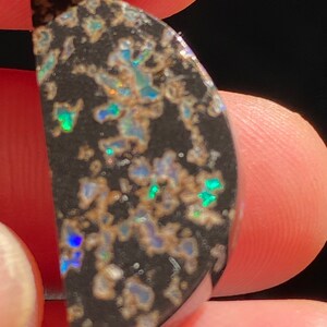 May include: A polished, semi-circular black opal gemstone with iridescent flashes of blue, green, and gold. The opal features a unique pattern of light-colored inclusions, creating a striking contrast against the dark background. The gemstone is held between fingers.