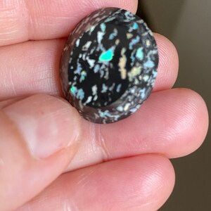 May include: Oval-shaped black opal gemstone with a speckled pattern of white, blue, and green inclusions. The polished surface reflects light, showcasing the play of color within the stone. A unique and eye-catching piece.