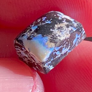 May include: A close-up of a rough, rectangular opal gemstone. The stone features a dark, almost black base color with streaks of white and flashes of blue and iridescent colors. The opal is held between two fingers.