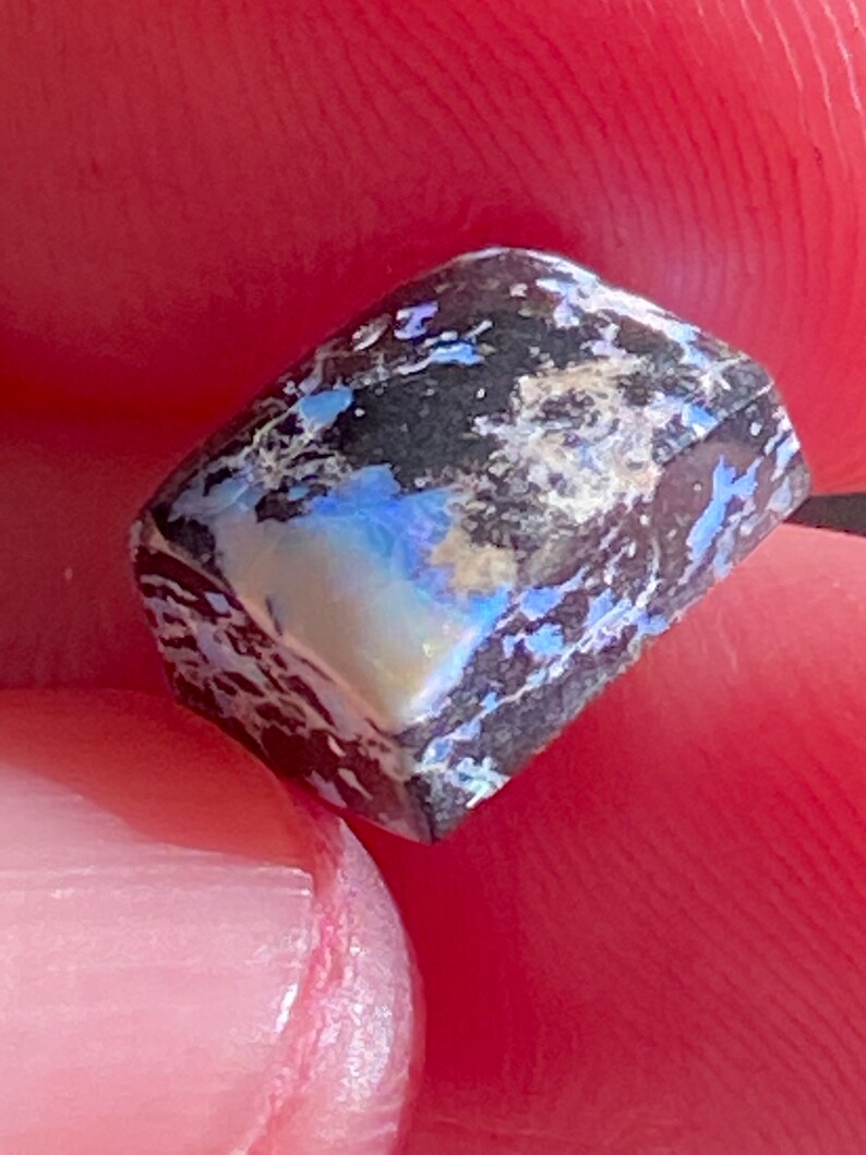 May include: A close-up of a rough, rectangular opal gemstone. The stone features a dark, almost black base color with streaks of white and flashes of blue and iridescent colors. The opal is held between two fingers.