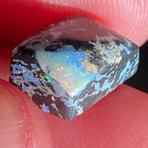 May include: A close-up of a rough-cut opal gemstone. The opal displays a vibrant play-of-color with flashes of blue, green, and red against a dark, textured background. The stone is held between fingers, showcasing its unique, natural beauty.