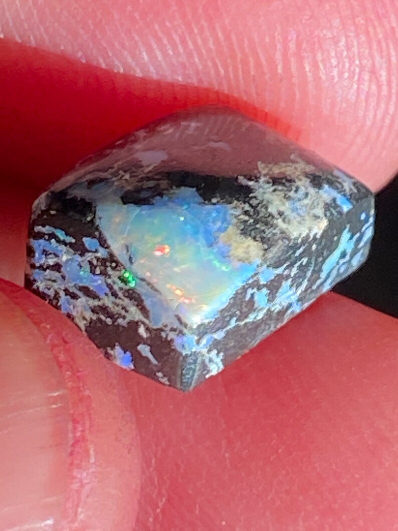 May include: A close-up of a rough-cut opal gemstone. The opal displays a vibrant play-of-color with flashes of blue, green, and red against a dark, textured background. The stone is held between fingers, showcasing its unique, natural beauty.