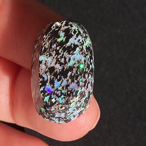 May include: A polished, oval-shaped gemstone with a black base and a mosaic of iridescent colors. The stone displays flashes of blue, green, and silver, creating a dynamic and eye-catching appearance. The gemstone is held in a hand.