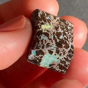 May include: A rough-cut opal gemstone with a dark brown matrix and vibrant flashes of color. The opal displays a mosaic pattern with areas of blue, green, and yellow. The stone is held between fingers, showcasing its natural beauty.