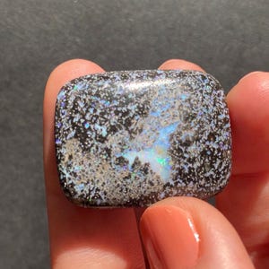 May include: A polished, square-shaped gemstone with a black base and a speckled pattern of silver, blue, and iridescent colors. The stone is held between fingers, showcasing its unique color play and texture. The gemstone is a natural opal.