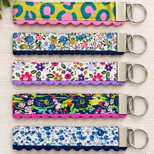 May include: Five fabric wristlet keychains with silver-tone metal hardware and split rings. Each strap features a unique pattern, including leopard print, floral designs, and botanical motifs, accented with decorative scalloped trim in various colours.