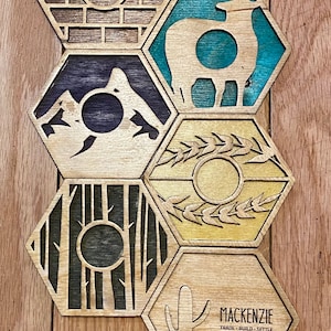 May include: Wooden hexagon tiles with laser-cut designs. The tiles feature images of a brick wall, a sheep, mountains, wheat, a forest, and a cactus. The bottom tile has the text "MACKENZIE TRADE BUILD SETTLE".