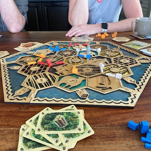 May include: A wooden board game with a blue and tan design, featuring hexagonal tiles and various game pieces in blue, orange, red, and white. Cards and other game components are scattered around the board on a wooden table.