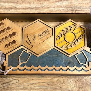May include: Wooden board game pieces in a wooden box. The pieces include hexagonal tiles with graphics and text, including a cactus, wheat, and the word "Mackenzie". The base has a blue water design.
