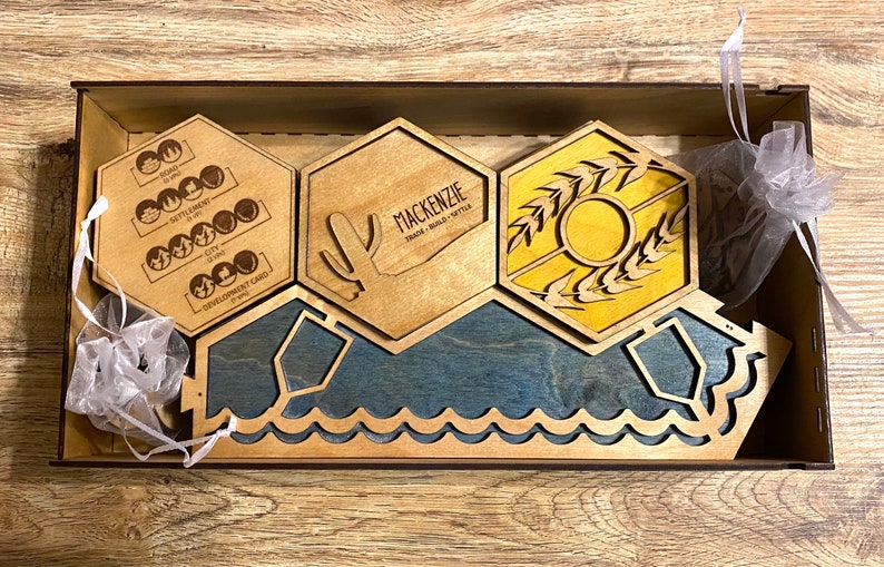 May include: Wooden board game pieces in a wooden box. The pieces include hexagonal tiles with graphics and text, including a cactus, wheat, and the word "Mackenzie". The base has a blue water design.