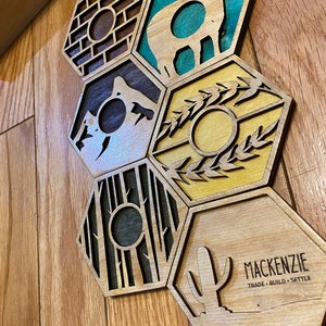 May include: Wooden hexagon game tiles with laser-cut designs. The tiles feature various landscape and object illustrations, including a mountain, trees, and a cactus. The text "MACKENZIE TRADE &bull; BUILD &bull; SETTLE" is visible on one tile. The tiles are arranged on a wooden surface.