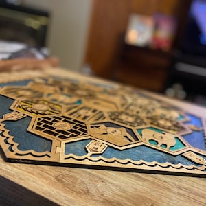 May include: Wooden board game with a hexagonal design. The game board features laser-cut wooden pieces with blue and green accents, depicting various terrains and game elements. The board is set on a wooden table.
