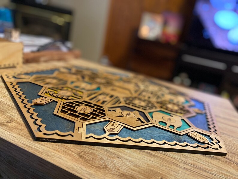 May include: Wooden board game with a hexagonal design. The game board features laser-cut wooden pieces with blue and green accents, depicting various terrains and game elements. The board is set on a wooden table.
