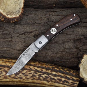 May include: A small pocket knife with a dark wood handle and a patterned steel blade. The blade is partially open and the knife is resting on a piece of wood with a natural texture. The knife has the words "Karve Ranch" engraved on the blade.