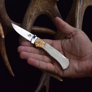 May include: A folding knife with a silver blade and a handle featuring a gold-colored, ornate design. The handle is light gray with small gold-colored rivets. The knife is held in a hand, with a dark background.