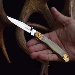 May include: A folding knife with a silver blade and a handle with a light gray and gold design. The blade has the text "Knives Ranch". The knife is held in a hand.