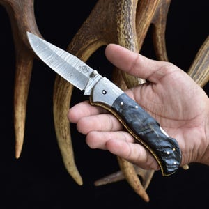 May include: A folding knife with a patterned steel blade and a dark, textured handle. The handle has a silver bolster and gold-colored accents. The knife is held in a hand against a backdrop of antlers.