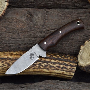 May include: A hunting knife with a brown wooden handle and a stainless steel blade with a Damascus pattern. The blade has a serrated edge and a small finger guard. The knife is resting on a piece of antler.