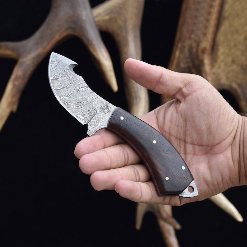 May include: A small, curved knife with a Damascus steel blade and a dark wood handle. The handle has a brass pin and a small hole at the end.