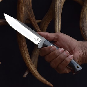 Puede incluir: Un cuchillo plateado con mango negro y filo dentado. El cuchillo se sostiene en una mano sobre un fondo de astas.
