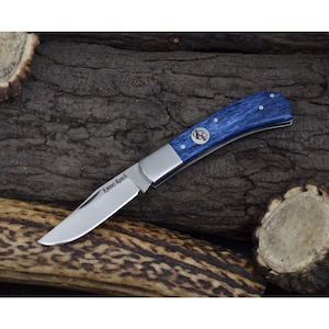Peut inclure: Un couteau de poche bleu et argent avec un manche en bois. La lame est partiellement ouverte et le couteau repose sur un morceau de bois. Le couteau porte les mots "Knives Ranch" gravés sur la lame.