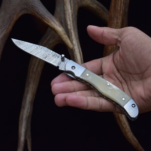 May include: A silver pocket knife with a white handle and a patterned blade. The knife is open and held in a hand.