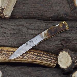 May include: A folding knife with a patterned blade and a handle featuring brown and cream tones. The knife has a silver-colored bolster and a decorative emblem near the blade's base. The handle appears to be made of a natural material.