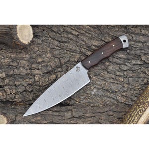 May include: A Damascus steel chef's knife with a dark wood handle. The knife has a textured blade and is resting on a piece of tree bark.