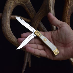 May include: A pocket knife with a partially open blade, featuring a cream-colored handle with gold-colored filigree and accents. The blade is silver with the text "Fence Ranch". The knife is held in a hand.