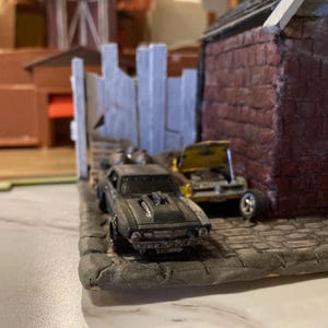 May include: A black and gray toy car with a detailed engine and a yellow toy car behind it. The cars are parked in front of a brick building with a wooden fence.