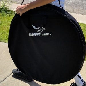 May include: A black circular carrying case with the text "MAYDAY GAMES" in white. The case has a handle and a logo featuring a stylized bird. The case is being carried by a person.