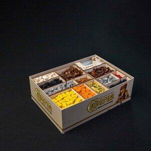 May include: A beige wooden game organizer with multiple compartments filled with various game pieces in colors like brown, yellow, orange, and gray. The box has the word "Caverna" printed on it.