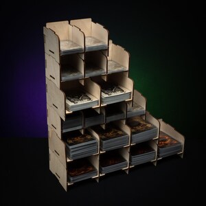May include: A wooden card organizer with multiple compartments arranged in a stepped design. Each compartment holds a stack of cards. The organizer is made of light-colored wood and is designed to store and display trading cards or board game cards.