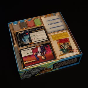 May include: A board game box with various game components. The box is blue with a light blue interior and contains cards, dividers, and other game pieces. The cards feature illustrations and text, including the words "Omnitron" and "Prime Wardens".
