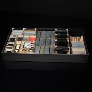 May include: A wooden game organizer with multiple compartments filled with game cards, tokens, and other game components. The organizer is rectangular and has a dark gray exterior with a patterned design. The interior features wooden dividers.
