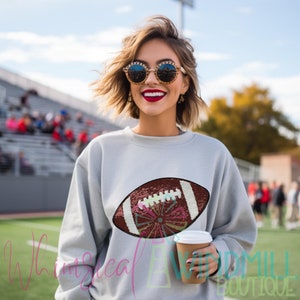 Op de afbeelding: Een grijs sweatshirt met een bruin en wit sequined voetbalontwerp. Het sweatshirt wordt gedragen door een persoon met kort blond haar en een zonnebril.