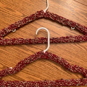 May include: Two crocheted hangers in a red and beige yarn. The hangers are made to fit over a standard plastic hanger.