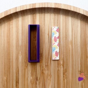 Peut inclure: Un moule rectangulaire en plastique violet et un morceau de papier rectangulaire à motifs sur une surface en bois.