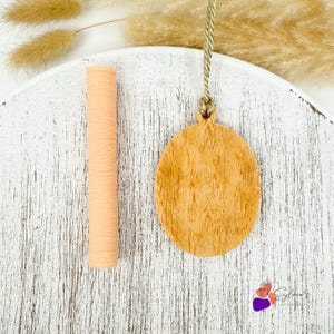 May include: A light peach-colored, textured rolling pin and a wooden oval ornament with a wood grain pattern. The ornament is suspended by a gold cord. The items are displayed on a white, distressed wooden surface.