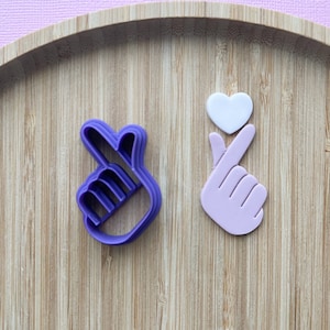 Puede incluir: Dos cortadores de galletas de plástico morado y rosa con forma de manos haciendo el signo del corazón coreano. La mano de la izquierda es un cortador, y la mano de la derecha es una forma sólida.