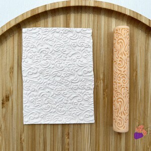 Può includere: Un mattarello in silicone bianco con un motivo floreale dettagliato e un foglio di silicone bianco con lo stesso motivo. Entrambi sono su una superficie di legno.