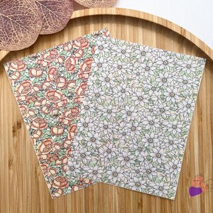 May include: Two patterned papers with floral designs. One features peach roses and green leaves, the other has white flowers with green leaves. The papers are on a wooden tray with decorative leaves.