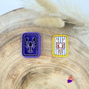 Puede incluir: Un cortador de galletas rectangular morado con diseño de langosta y borde elevado, junto a una galleta rectangular a juego con fondo de rayas blancas y azules, una ilustración de langosta roja y un borde amarillo con pequeñas líneas.
