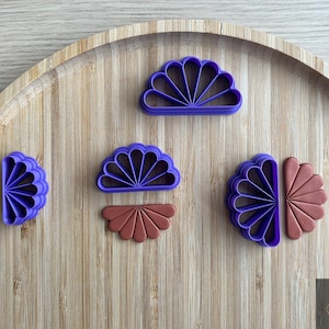 Puede incluir: Cinco cortadores de galletas de pl&aacute;stico morado en forma de abanicos, uno de ellos de color marr&oacute;n. Los cortadores est&aacute;n dispuestos sobre una superficie de madera.