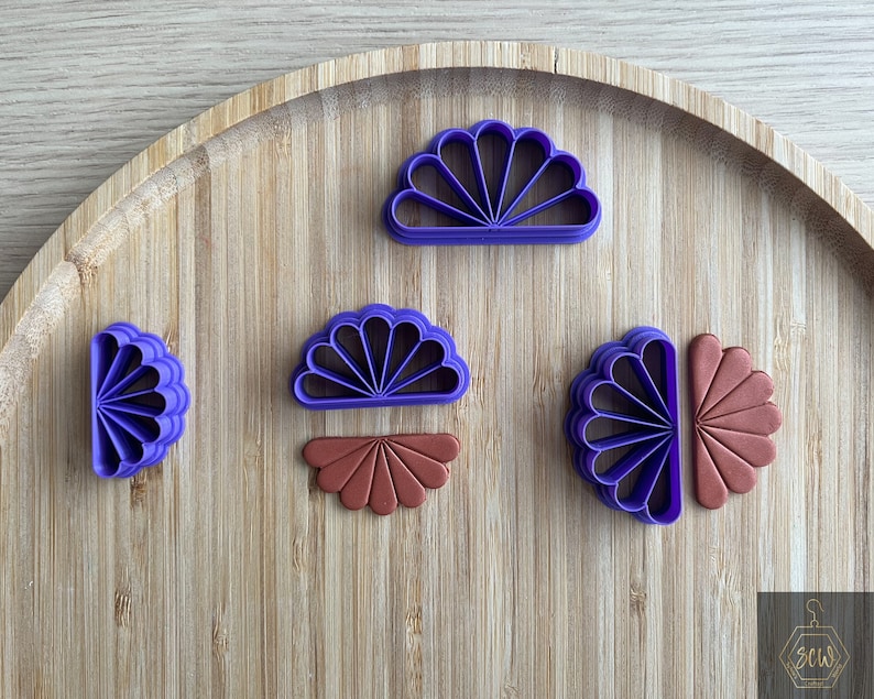 Puede incluir: Cinco cortadores de galletas de pl&aacute;stico morado en forma de abanicos, uno de ellos de color marr&oacute;n. Los cortadores est&aacute;n dispuestos sobre una superficie de madera.