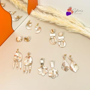 May include: Assortment of handmade polymer clay earrings in various shapes and colors, including stars, moons, flowers, and abstract designs. The earrings are displayed on a light beige surface.