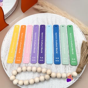 Könnte beinhalten: Ein Satz von acht bunten Holzlinealen mit dem Text "Sylvia's Crafted World" und Messungen in Millimetern. Die Lineale sind in einem Regenbogenmuster von Gelb bis Grün angeordnet.