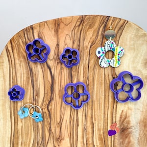 Puede incluir: Una tabla de cortar de madera con cinco cortadores de galletas con forma de flor morada y un pendiente con estampado floral colorido.
